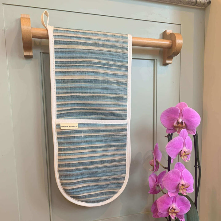 double oven glove with blue and sandy stripes Hanging over a wooden bar off a kitchen cupboard door with a pink flower