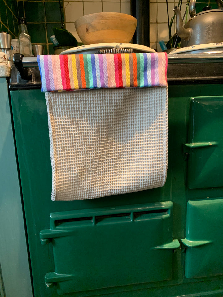 Roller towel with rainbow header and beige cotton waffle fabric hanging over a green Aga range cooker front railing 