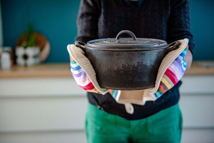 person with green trouser and charcoal jumper holding a cast iron black dish with a pair of rainbow double oven gloves, kitchen units and blue backdrop