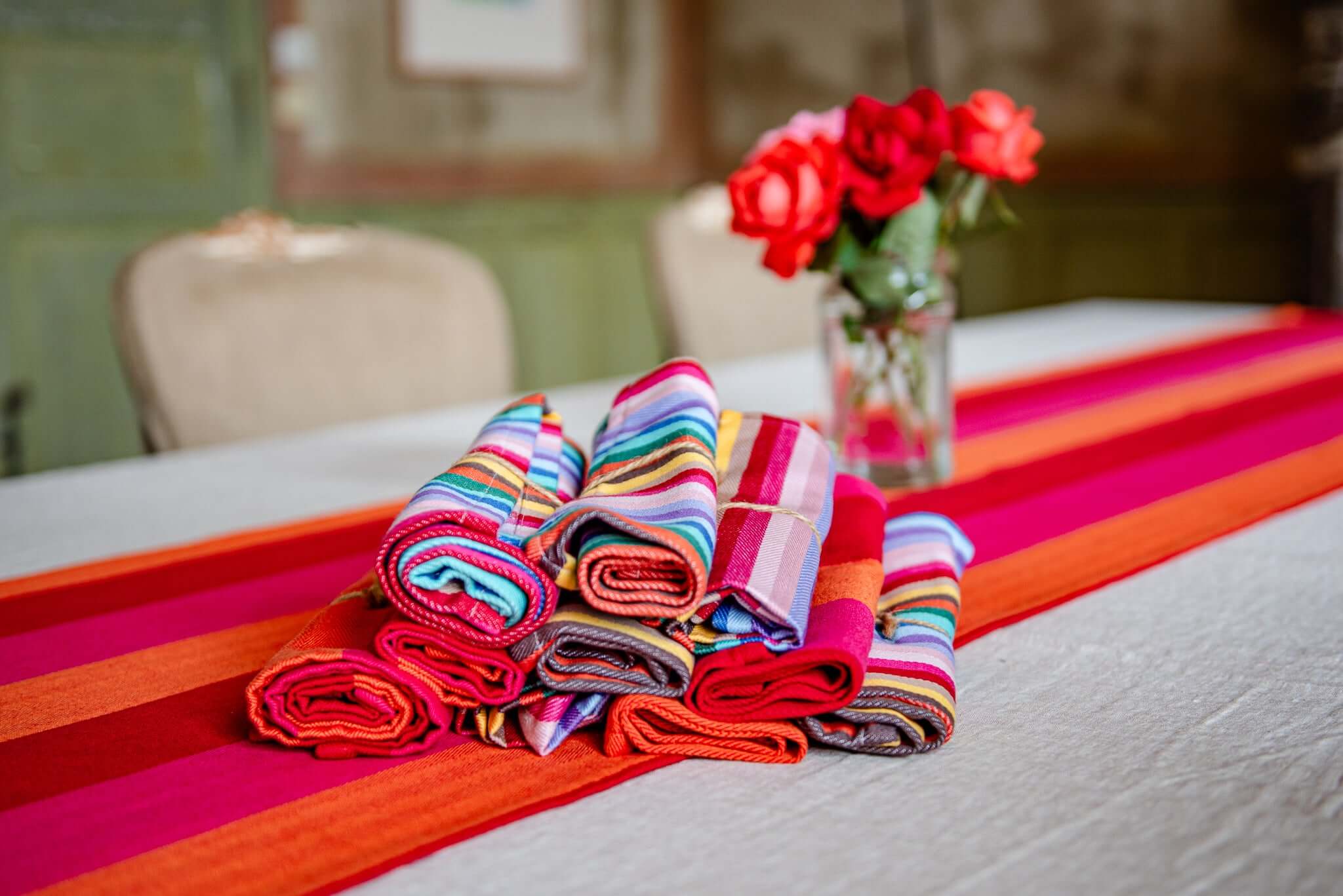 red orange pink table runner on a ecru linen table cover with a pile of rolled up napkins rainbow and matching red orange pink in focus. A vase with red roses and chairs in the background