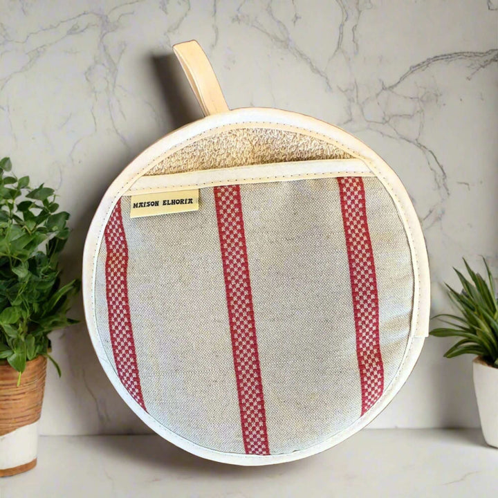  round oven mitt, pot holder type linen with red french style stripes on a white marble kitchen top and backdrop with green plants on either side