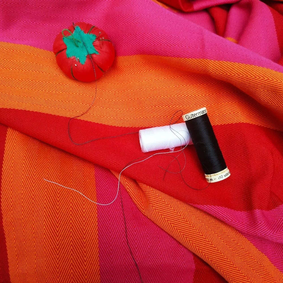 Close up of red orange and pink herringbone fabric with a white and a black cotton bobbin and a tomatoed shaped needles holder 