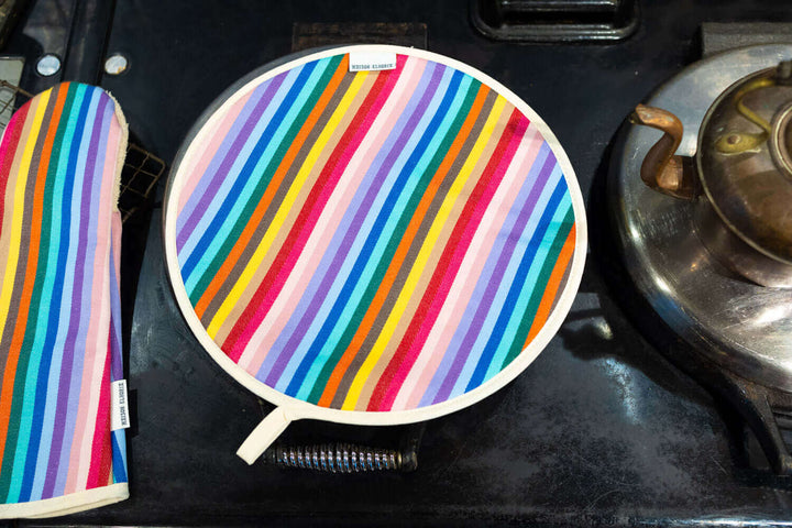close up shot of a single rainbow chef pad on an Aga range cooker top hob , showing part of an antique kettle to the right hand side and part of a matching rainbow long oven glove gauntlet style to the left hand side