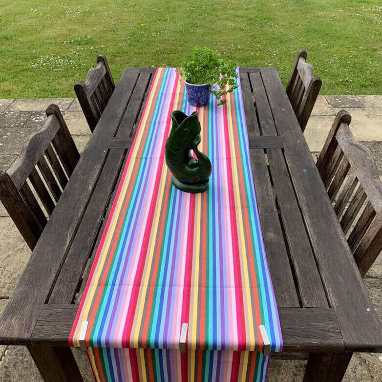 Rainbow organic and recycled cotton table runner on a garden table outside with 4 chairs on a terrasse with grass in the background a fish shaped vase and a pot of parsley on top of it