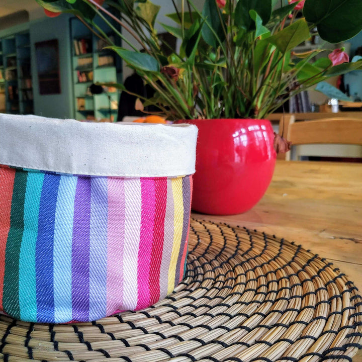Rainbow cotton round storage pot bag on a place matt on wooden table with a red plant pot behind it