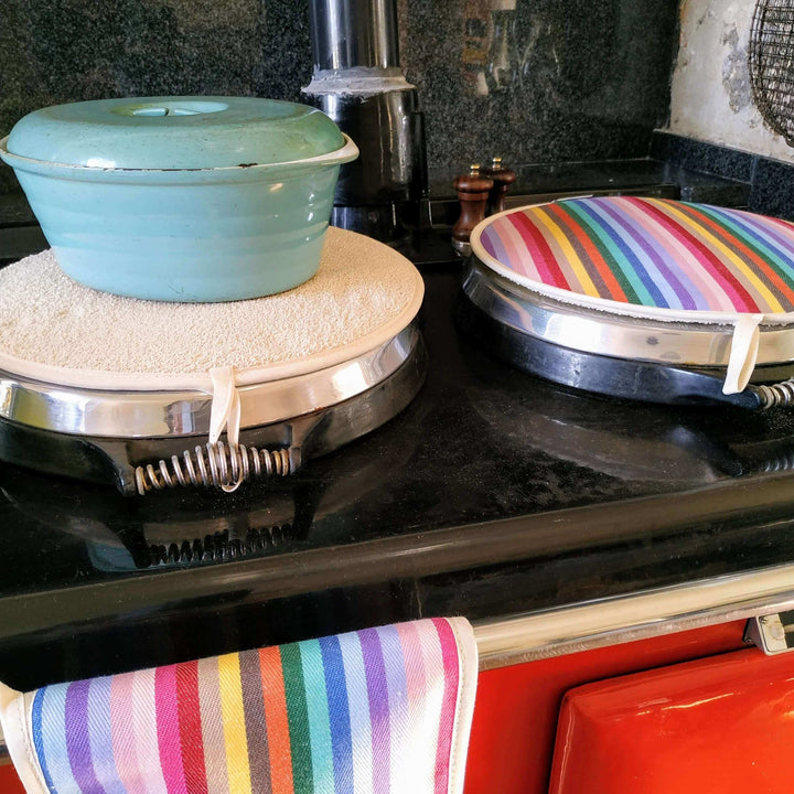 Two Rainbow premium quality insulating Aga hob plate cover on a red aga . One aga cover is upside down with a cast iron turquoise pot on it. A rainbow tea towel hangs on the front of the aga