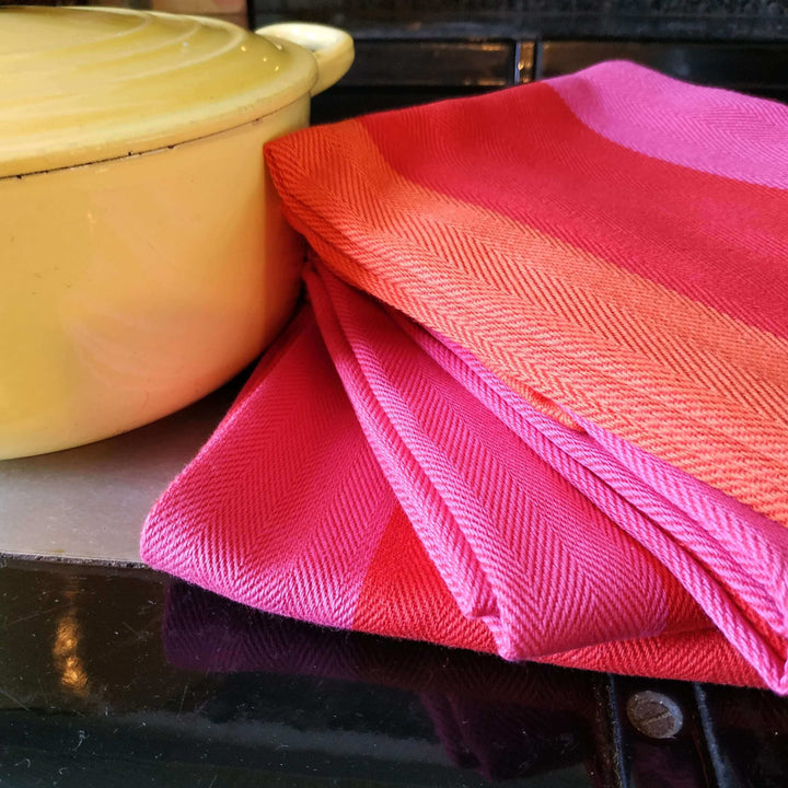 Three red orange pink striped tea towels folded on top of each other with a yellow antique Le Creuset dish beside
