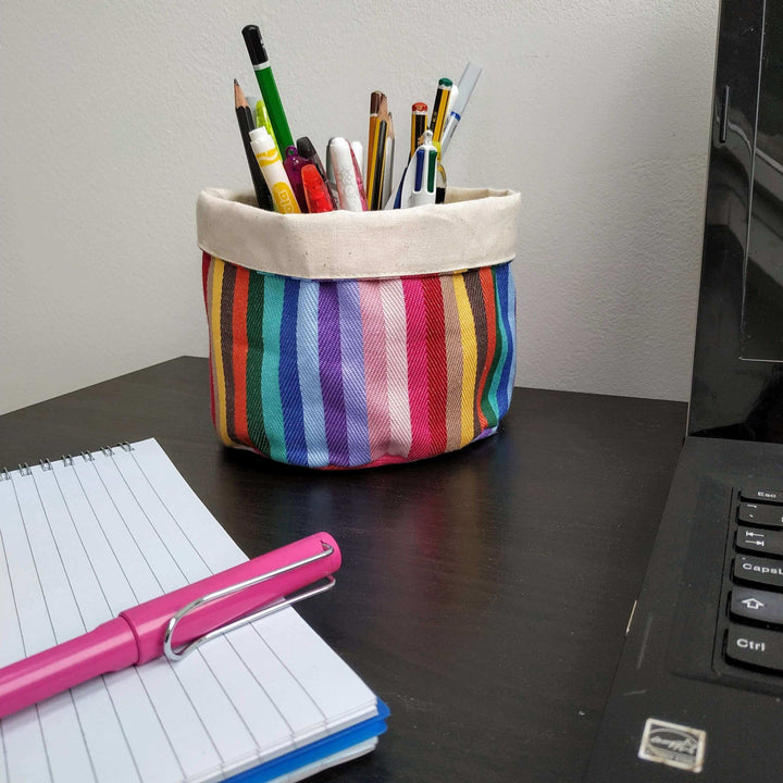rainbow cotton round storage pot bag, holding pencils, on a black desk with a notepad and a pink pen on it.