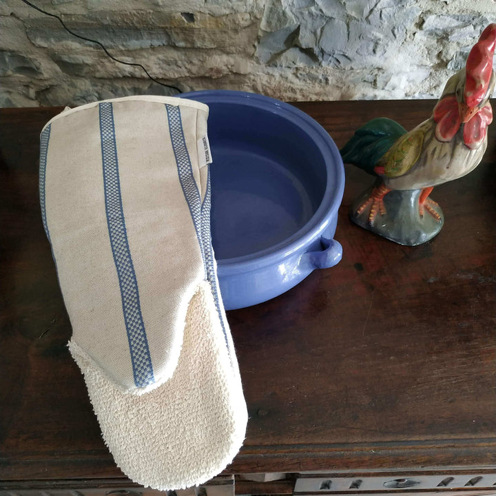 Gauntlet long oven glove line union with blue stripes above an oven ware blue dish  on a wooden dresser with a cockerel statuette  with a stone wall in the back