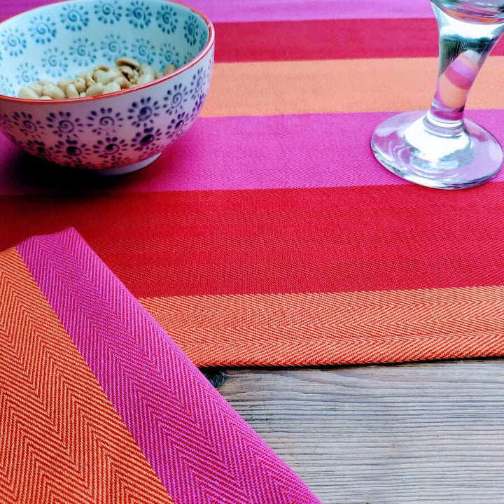 cut image of a table runner and napkin red orange pink organic and recycled cotton fabric on a wooden table with a glass and a bowl of peanuts