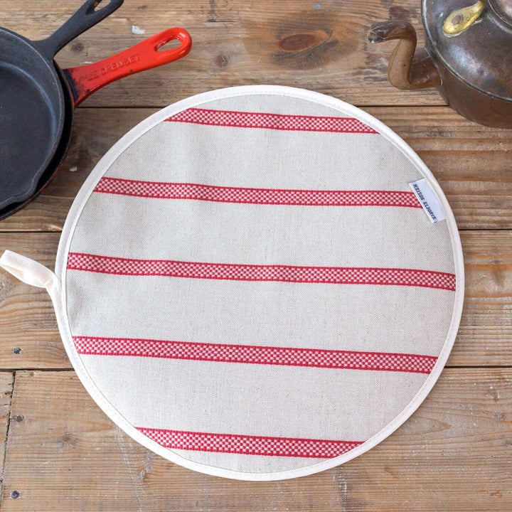 Linen union with red stripes insulating Aga hob plate cover on wooden surface with cast iron pans and antique kettle
