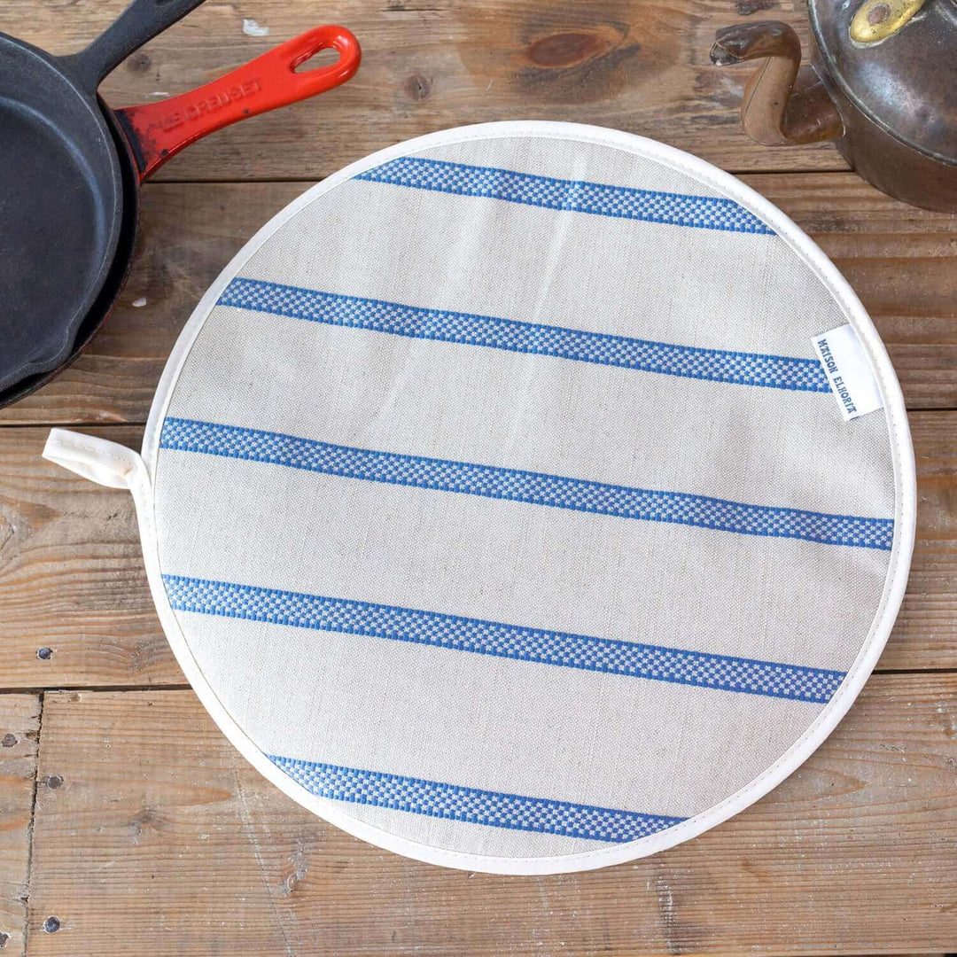 Linen union with blue stripes insulating Aga hob plate cover on wooden surface with cast iron pans and antique kettle